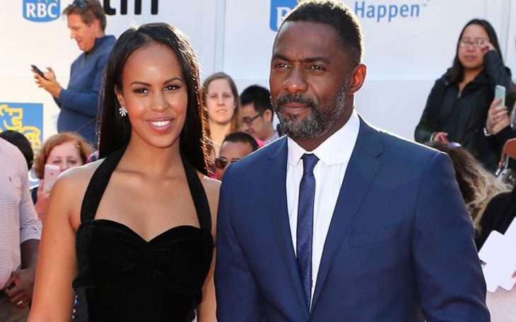 Idris Elba y Sabrina Dhowre, mujer afroamericana con vestido negro sonriendo y hombre afroamericano con traje azul y rostro serio