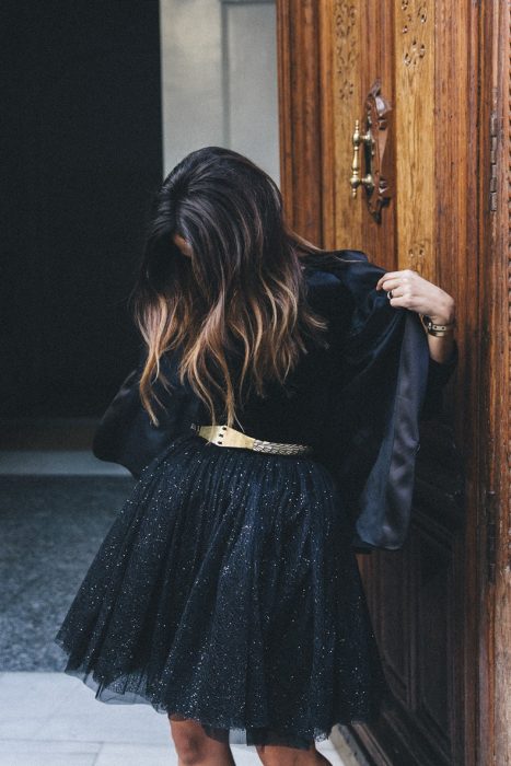 Chica de cabello castaño con luces usando vestido negro con brillos, cinto dorado y chaqueta negra