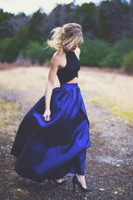 Chica de cabello rubio recogido en una coleta usando un top negro con una falda larga de satín azul rey y zapatillas negras en un prado