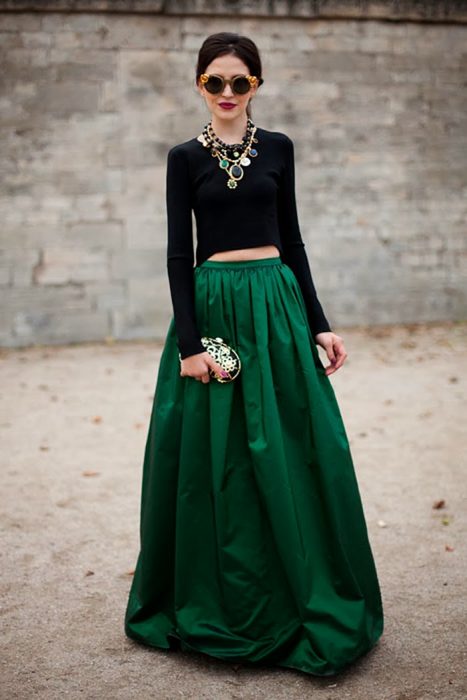 Chica con el cabello recogido usando lentes redondos, collar grande, blusa negra de manga larga y una falda larga de satín verde esmeralda