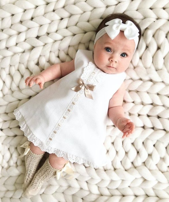 Bebé niña recostada en una manta tejida, usando un vestido de color blanco con un moñito blanco y calcetitas doradas 