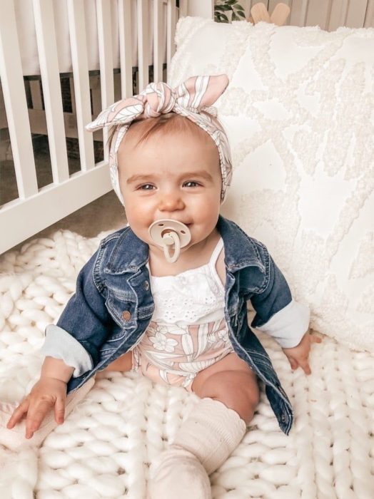 Bebé niña sentada en su cuna usando una camisetita de mezclilla con una bandita en la cabeza 