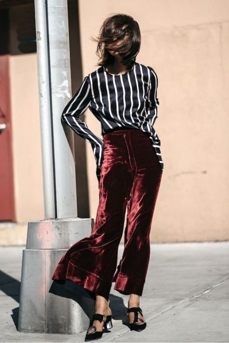 Chica con el rostro tapado por el cabello, con blusa de rayas blancas y negras usando pantalón de terciopelo rojo acampanado y zapatillas abiertas