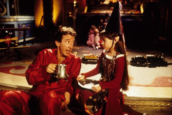 Hombre vestido de rojo tomando una bebida caliente con una elfa con vestido rojo y gorro de Navidad