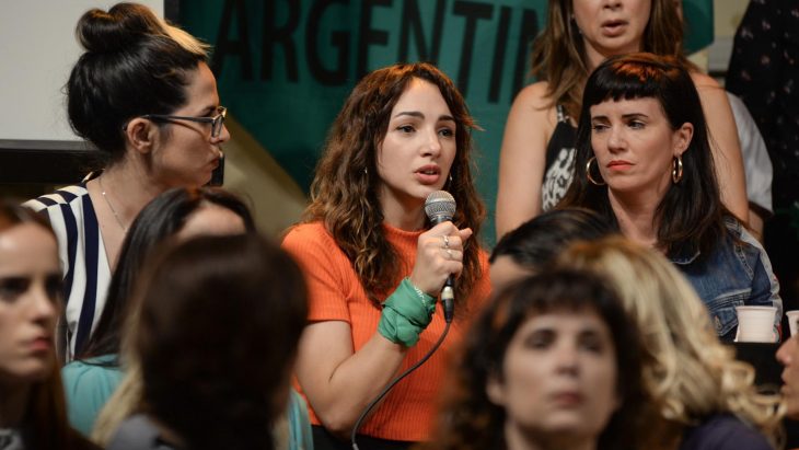 Thelma Fardín, mujer de blusa anaranjada y cabello chino hablando por un micrófono rodeada de más mujeres