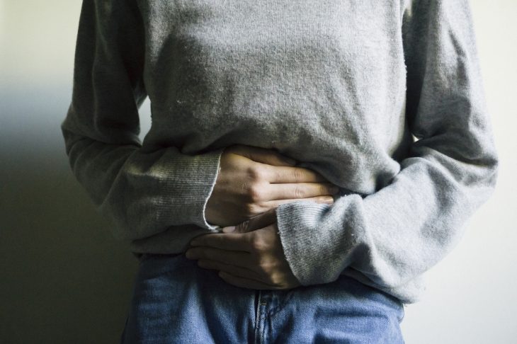 Mujer con cólicos, suéter gris y con las manos sobre el abdomen