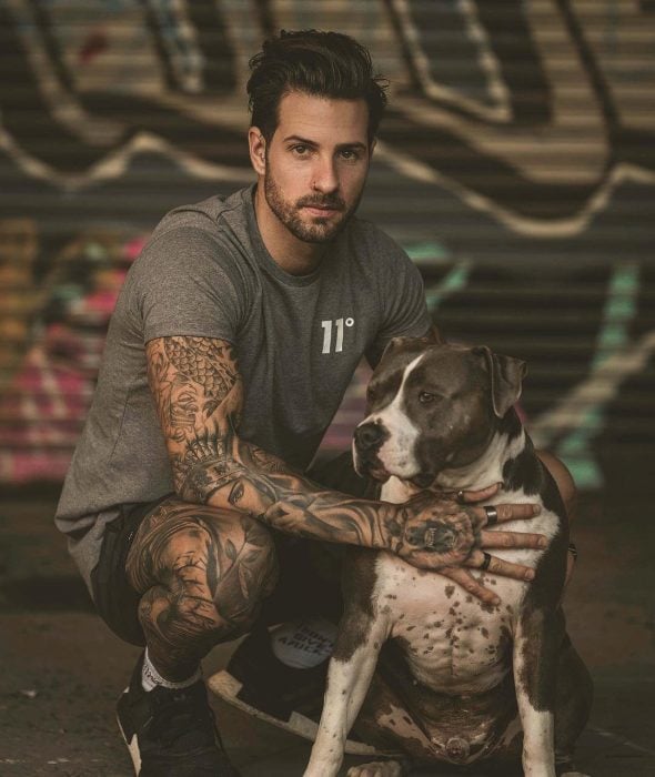 Hombre con tatuajes en los brazos y piernas a lado de un perro raza pitbull