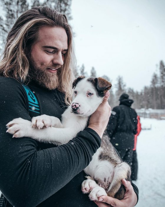 Hombre rubio de cabello largo y barba cargando un perro blanco con manchas café