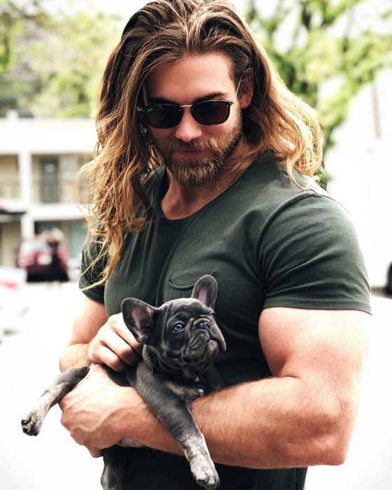 Hombre musculoso de cabello largo y barba cargando a un perro bulldog francés negro