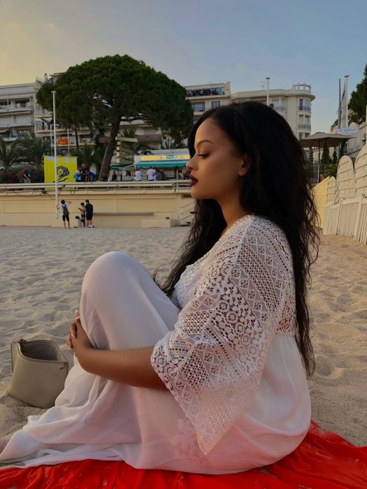 Mujer afroamericana con cabello largo y vestido blanco sentada en la playa que se parece a Rihanna