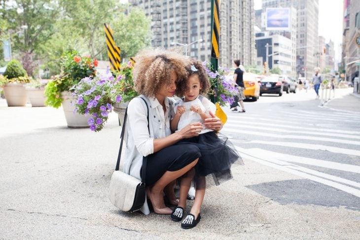 Mamá e hija de cabello rizado, usando cada quien una falda negra, chamarra de mezclilla, paradas en medio de la avenida, posando para una foto familiar