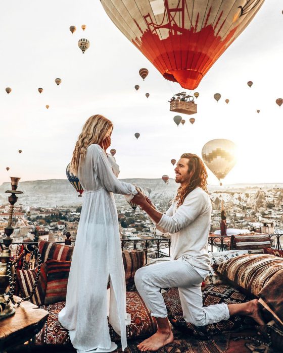 Novio de cabello largo hincado pidiéndole matrimonio a su novia de cabello rubio, ambos vestidos de blanco con globos aerostáticos en el fondo