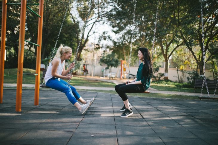 Dos chicas en un parque, sentadas en un columpio, comiendo helado, charlando, al atardecer