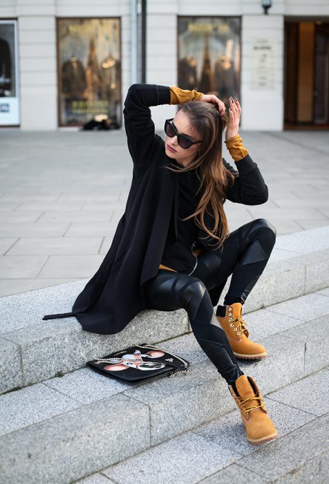 Chica sentada en escaleras peinando su cabello, trae unos leggins negros, blusa negra y suéter negro. Además de botas militares cafés y gafas en el rostro