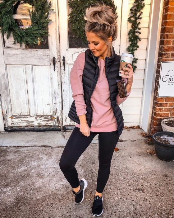 Chica posando enfrente de una puerta de madera blanca rustica, con leggins negro, suéter rosa con chaleco azul. De calzado lleva tenis negros y en su mano un café caliente
