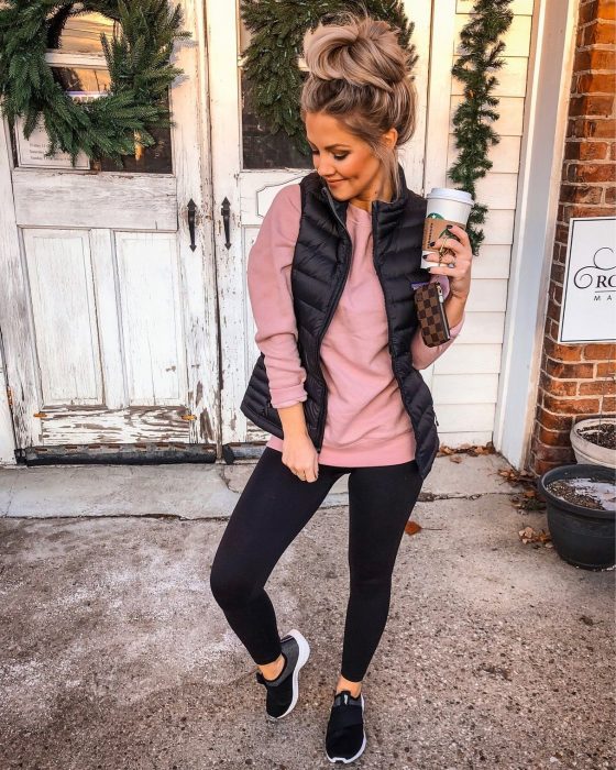 Chica posando enfrente de una puerta de madera blanca rustica, con leggins negro, suéter rosa con chaleco azul. De calzado lleva tenis negros y en su mano un café caliente