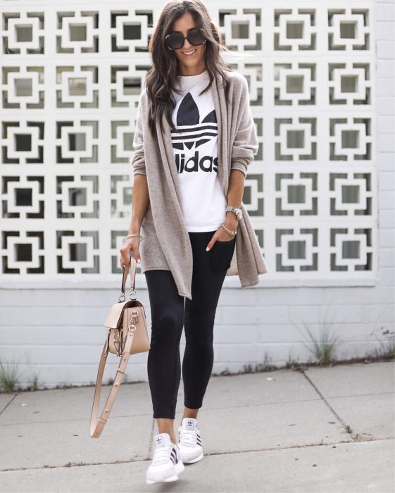 Chica posando enfrente de una pared con leggins negros, camisa blanca y estampado de addidas, con tenis blancos