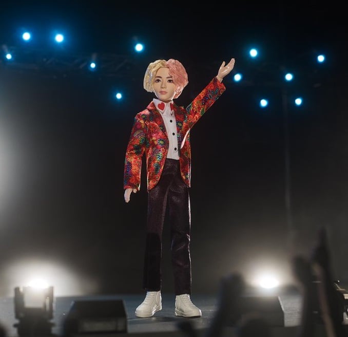 Muñeco inspirado en el cantante de la banda de K-pop, BTS, V, usando un saco de color rojo con pantalones de color café, tenis y camisa blanca 