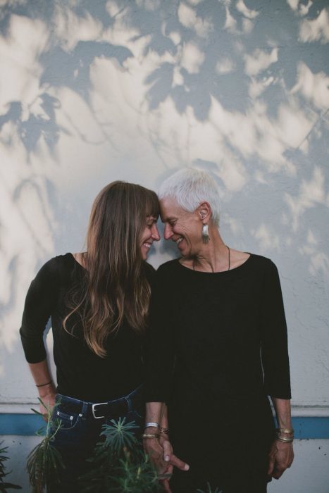 Madre de cabello blanco y corto e hija de cabello castaño con flequillo tomadas de la mano y sonriendo con los ojos cerrados