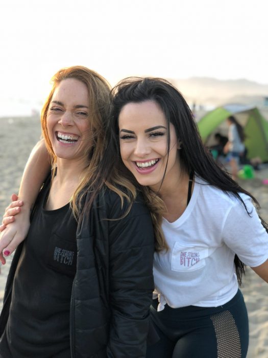 Madre de cabello rubio e hija de cabello castaño en la playa sonriendo mientras se abrazan