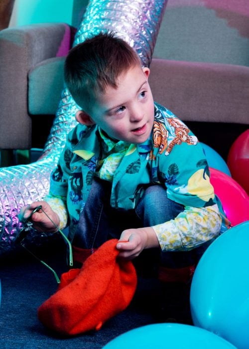 Niño modelo con síndrome de Down guardando unas gafas oscuras en un saco de tela rojo en una sala llena de globos 