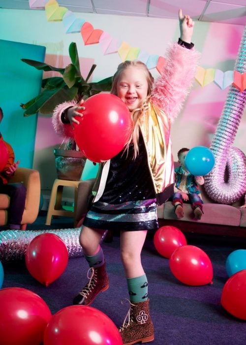 niña modelo con síndrome de Down jugando en una sala de espera con globos rojos, zalzando su mano izquierda para señalar el techo y sosteniendo un globo con su mano derecha