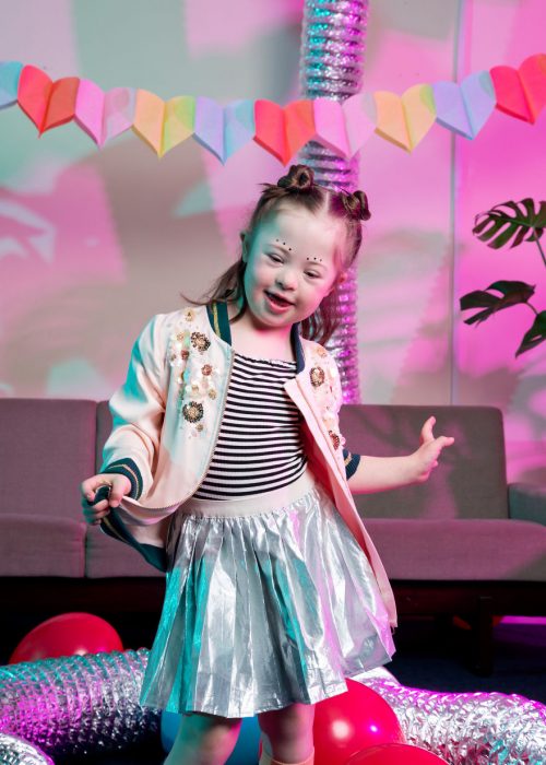 niña modelo con síndrome de Down bailando en una sala de espera llena de globos rojos y decorada con corazones de papel de colores