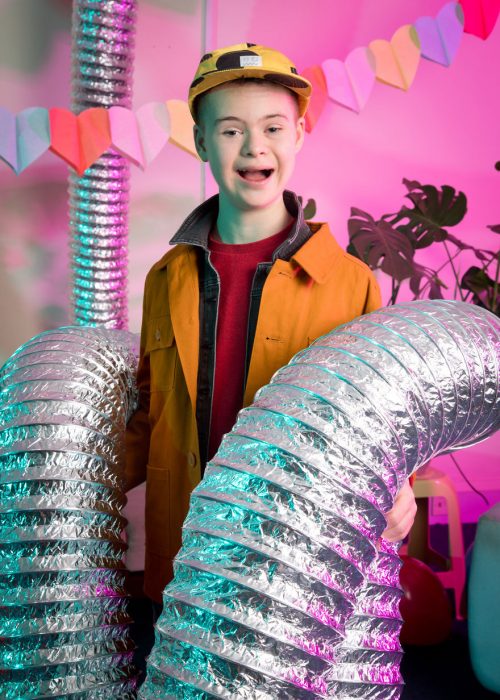 niño modelo co síndrome de Down sosteniendo un par de tubos metálicos ligeros, sonriendo, usando chaqueta y gorra amarillas