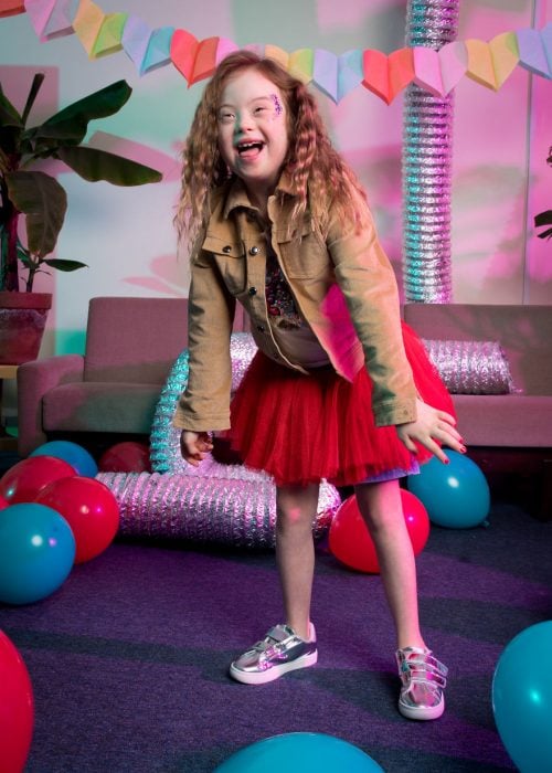 niña con síndrome Down modelando falda de tutu en color rojo, usando tenis y chqueta café de pana, sonriendo, dentro de una sala de espera decorada con globos en azul y rojo 