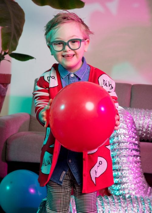 niño modelo con síndrome de Down sosteniendo un globo rojo entre sus manos, llevando gafas de marco gris, suéter rojo, camisa denim. pantalón a cuadros