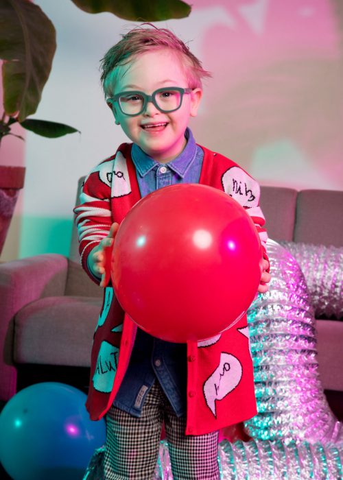 niño modelo con síndrome de Down sosteniendo un globo rojo entre sus manos, llevando gafas de marco gris, suéter rojo, camisa denim. pantalón a cuadros