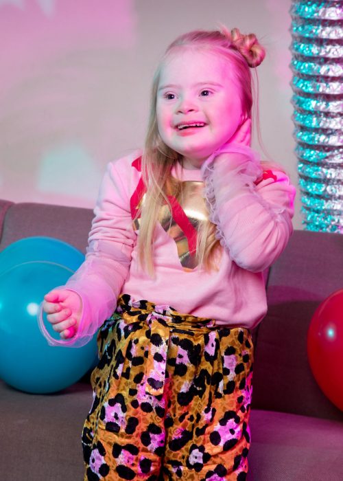 niña modelo con síndrome de Down acariciando su cabello rubio, sonriendo de manera nerviosa, llevando pantalón flojo con decorado en manchas y sudadera rosa, parada frente a un sofá con globos rojos y azul