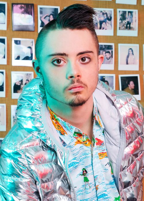  Modelo con síndrome de Down usando chaqueta metálica, camisa hawaiana, con ojos delineados, modelando dentro de una oficina decorada con fotografías antiguas