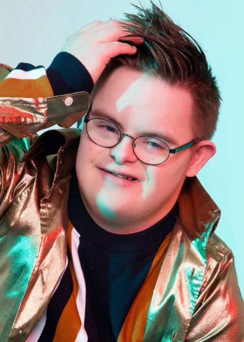 Modelo con síndrome Down usando anteojos, chaqueta metálica, acariciando su cabello ligeramente con la mano izquierda y sonriendo para una fotografía
