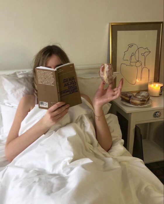 Mujer leyendo en la cama y comiendo dona