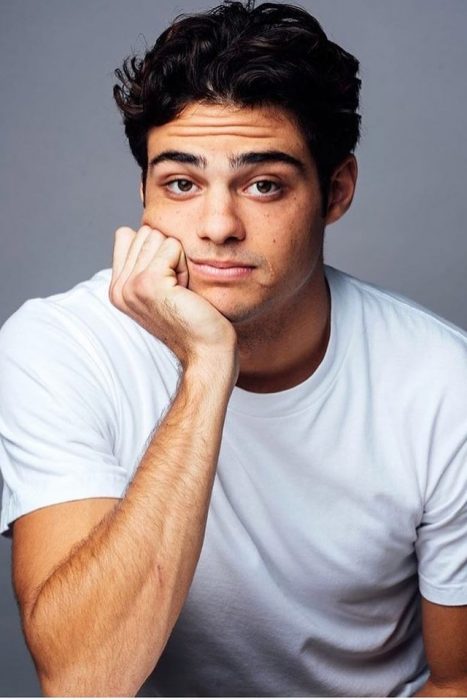 Noah Centineo usando camisa blanca, recargando su barbilla en su mano derecha