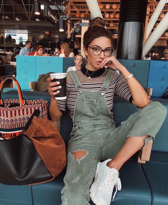 Chica con overol de mezclilla verde y camisa a rayas tomando un café