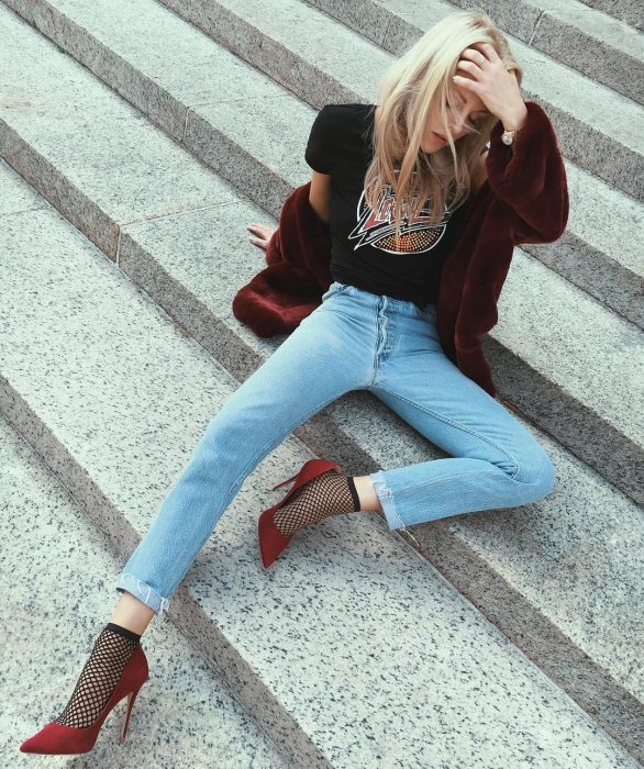 Chica rubia con blusa rockera, abrigo rojo vino afelpado, jeans y tacones rojos con medias de red