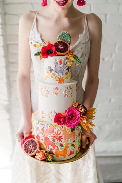 Mujer sosteniendo un pastel de tres pisos con betún de vainilla blanco y decorado con flores naturales de colores