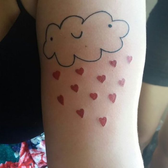 Tatuaje en el brazo mal hecho y feo de una nube sonriente que arroja lluvia de corazones