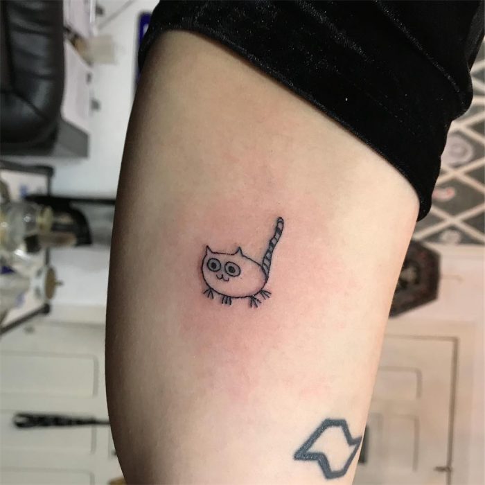 Tatuaje en la pierna como dibujo infantil de un gato gordo de patas cortas hecho por la tatuadora Helen Fernandes