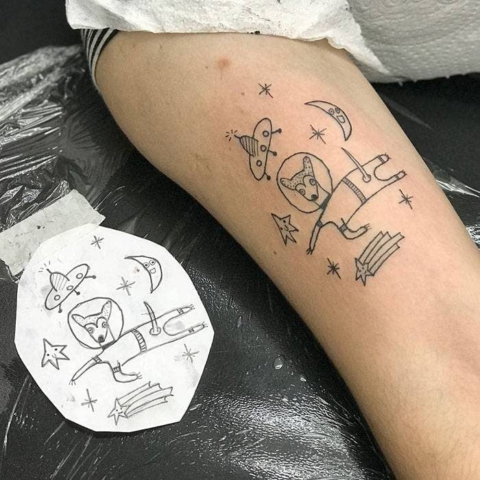 Tatuaje en el brazo parece dibujo infantil de un perro astronauta en el espacio con estrellas fugaces y ovnis hecho por la tatuadora Helen Fernandes