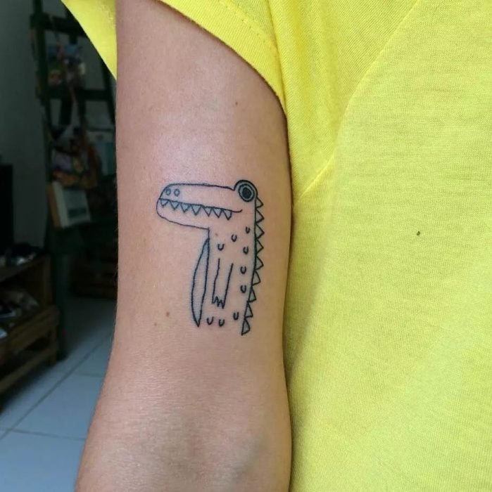 Tatuaje en el brazo parece dibujo de niño de un cocodrilo por la tatuadora Helen Fernandes