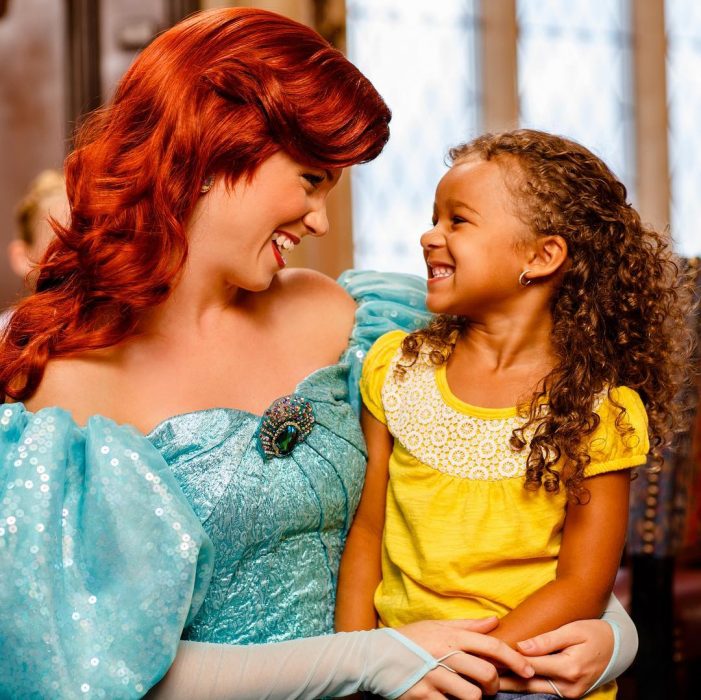 Mujer vestida de Ariel de la Sirenita con una niña sentada en sus piernas mientras ambas sonríen 