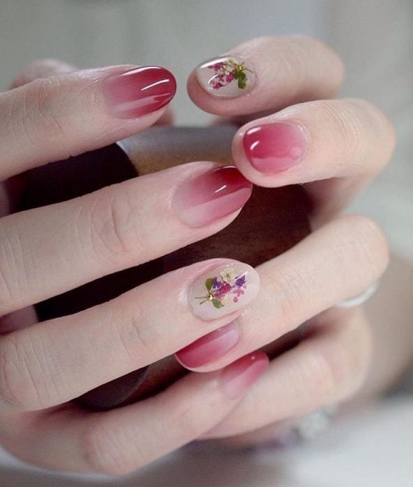 Manos de mujer con uñas pintadas con esmalte rosa degradado y pequeñas flores blancas, rosas y moradas con una piedra dorada