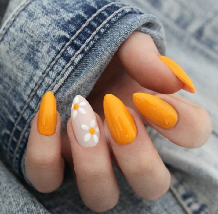 Mano de mujer con uñas largas en forma de almendra con una uñas pintada con flores blancas, margaritas, y las demás de color amarillo anaranjado para primavera