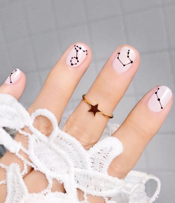 Manicura femenina y minimalista de constelaciones con esmalte color perla y estrellas negras con un anillo dorado de estrella