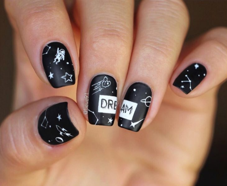 Manicura creativa y divertida de constelaciones, con esmalte negro y estrellas, cohetes, ateroides y astronauta blancos