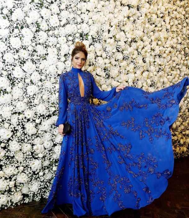 Chica con vestido de fiesta largo color azul royal con mangas y pedrería frente a una pared de flores blancas