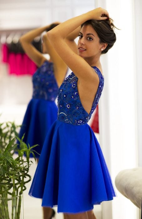 Chica de cabello castaño frente a un espejo con vestido de fiesta corto color azul royal con ornamentos y pedrería en la parte superior y sin mangas